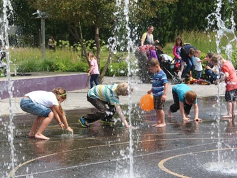 Kinder spielen begeistert im spritzenden Wasser eines Brunnens in einem grünen Park bei Sonnenschein.