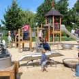 Kinder spielen auf einem Spielplatz mit Kletterturm und Sandfläche, umgeben von Bäumen und Erwachsenen.