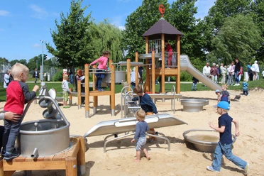 Wasserspielplatz zwischen Ober- und Untersee Kinder spielen auf einem Spielplatz mit Kletterturm und Sandfläche, umgeben von Bäumen und Erwachsenen.