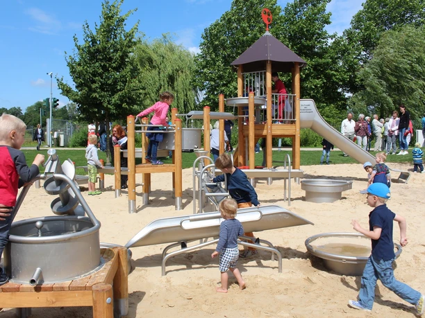 Wasserspielplatz zwischen Ober- und Untersee Kinder spielen auf einem Spielplatz mit Kletterturm und Sandfläche, umgeben von Bäumen und Erwachsenen.