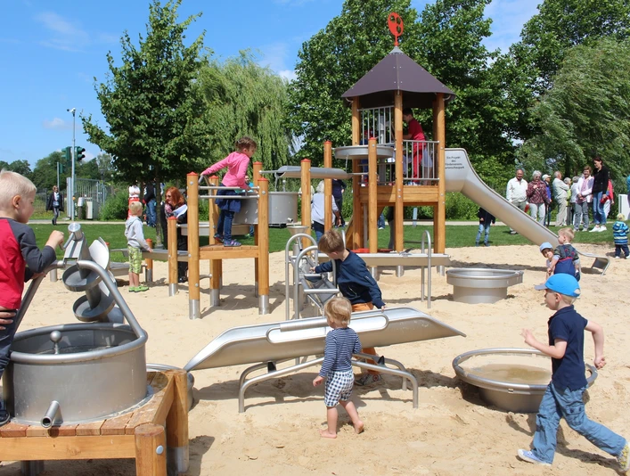 Wasserspielplatz zwischen Ober- und Untersee Kinder spielen auf einem Spielplatz mit Kletterturm und Sandfläche, umgeben von Bäumen und Erwachsenen.