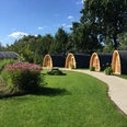 Drei hölzerne Campingpods mit Bogendach stehen auf einer grünen Wiese entlang eines gepflasterten Pfads.