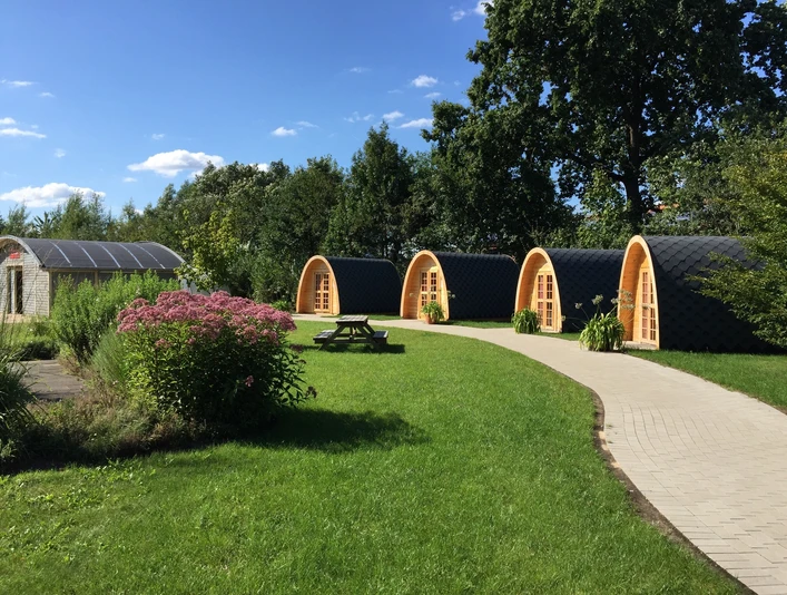 Campingpods - urige Übernachtungsmöglichkeit Drei hölzerne Campingpods mit Bogendach stehen auf einer grünen Wiese entlang eines gepflasterten Pfads.