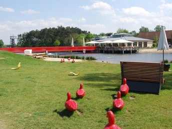 Grüne Wiese mit roten Kunstobjekten, im Hintergrund ein See und Gebäude mit Terrasse.