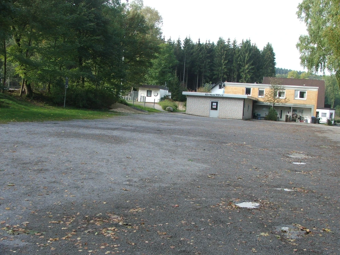 Parkplatz Freibad Großer, leerer Parkplatz vor einem Freibad mit umliegenden Bäumen und einem Gebäude im Hintergrund.