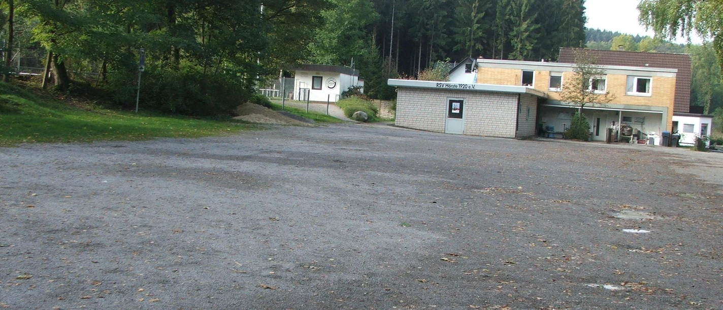 Parkplatz Freibad Großer, leerer Parkplatz vor einem Freibad mit umliegenden Bäumen und einem Gebäude im Hintergrund.