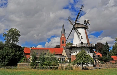 Klostermühle Lahde Historische Windmühle neben einer Kirche, umgeben von Bäumen und einem leichten Wolkenhimmel.