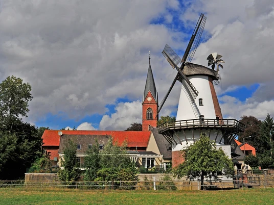Klostermühle Lahde Historische Windmühle neben einer Kirche, umgeben von Bäumen und einem leichten Wolkenhimmel.