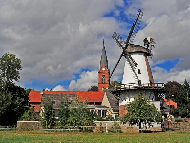 Klostermühle Lahde Historische Windmühle neben einer Kirche, umgeben von Bäumen und einem leichten Wolkenhimmel.