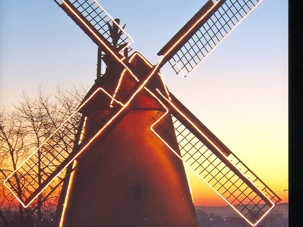 Eine historische Windmühle bei Sonnenuntergang, beleuchtet von Lichterketten mit Bäumen im Hintergrund.