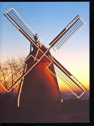 Valentins Mühle Eine historische Windmühle bei Sonnenuntergang, beleuchtet von Lichterketten mit Bäumen im Hintergrund.