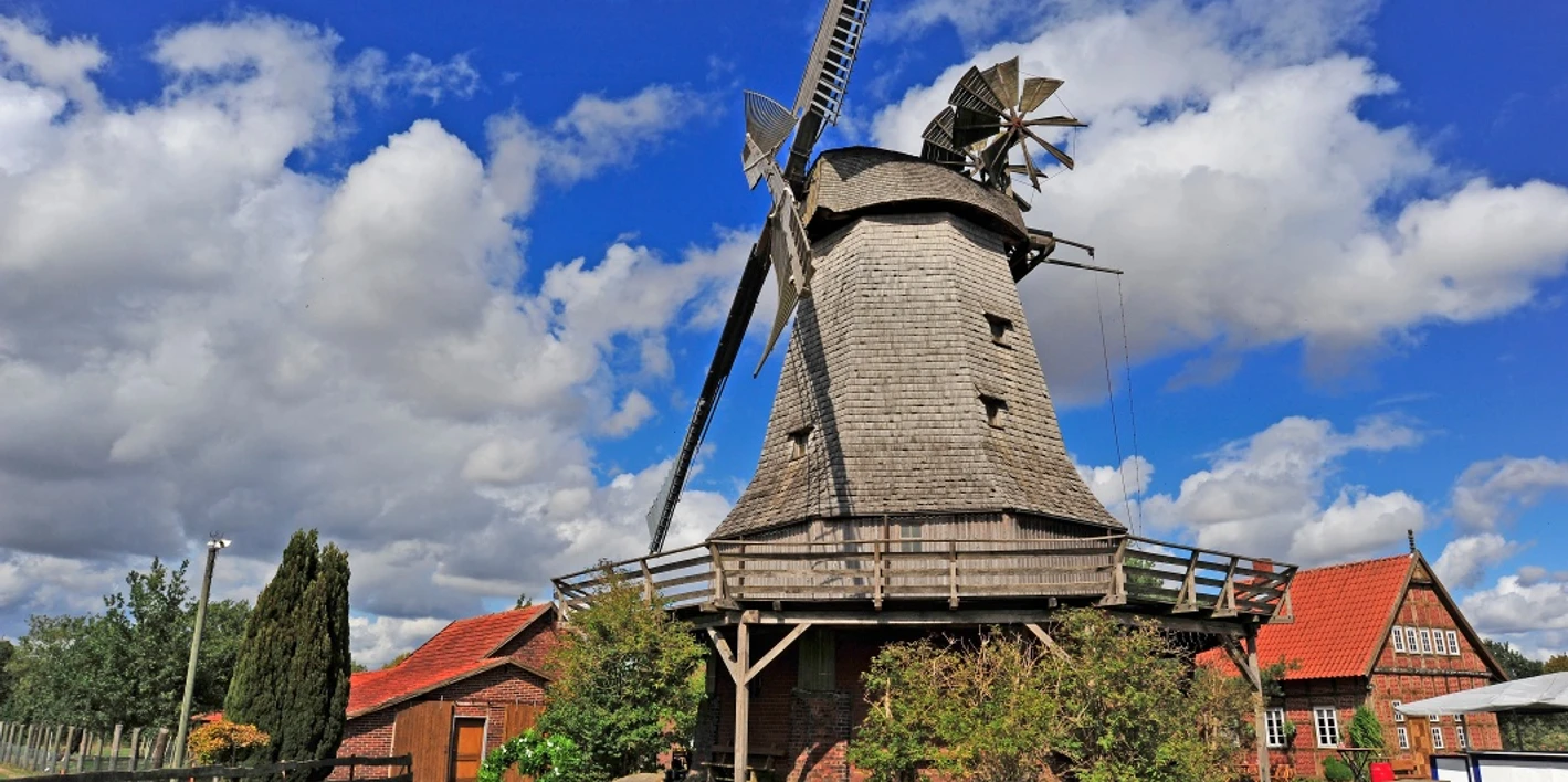 Meßlinger Mühle 2 Historische Windmühle mit grauen Schindeln, Holzgalerie und Flügeln vor blauem Himmel und Wolken.