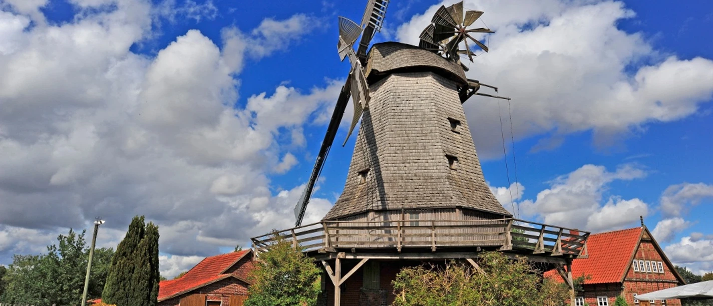 Meßlinger Mühle 2 Historische Windmühle mit grauen Schindeln, Holzgalerie und Flügeln vor blauem Himmel und Wolken.