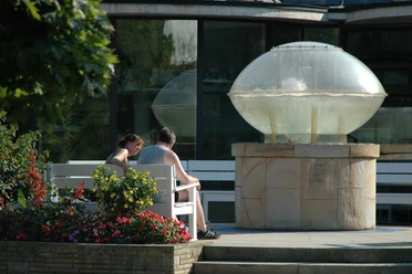 Thermalsprudel III Ein Paar sitzt auf einer Bank neben einem modernen Brunnen mit einer Glaskuppel in einem Garten.