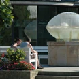 Thermalsprudel III Ein Paar sitzt auf einer Bank neben einem modernen Brunnen mit einer Glaskuppel in einem Garten.