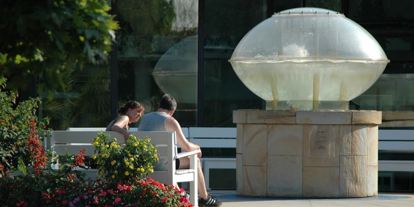 Thermalsprudel III Ein Paar sitzt auf einer Bank neben einem modernen Brunnen mit einer Glaskuppel in einem Garten.