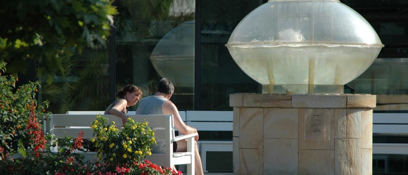Thermalsprudel III Ein Paar sitzt auf einer Bank neben einem modernen Brunnen mit einer Glaskuppel in einem Garten.