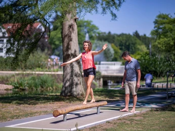 Kneipp-Pfad im Erlebnis- & Gesundheitspark Bad Salzuflen Frau balanciert barfuß auf einem Holzstamm, während ein Mann zusieht, in einem grünen Parkumfeld.