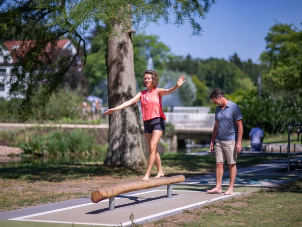 Kneipp-Pfad im Erlebnis- & Gesundheitspark Bad Salzuflen Frau balanciert barfuß auf einem Holzstamm, während ein Mann zusieht, in einem grünen Parkumfeld.