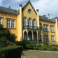 Gelbes Schloss im neugotischen Stil mit Spitzbogengiebel und verziertem Balkon in grüner Umgebung.