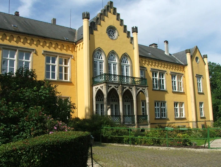 Gelbes Schloss im neugotischen Stil mit Spitzbogengiebel und verziertem Balkon in grüner Umgebung.