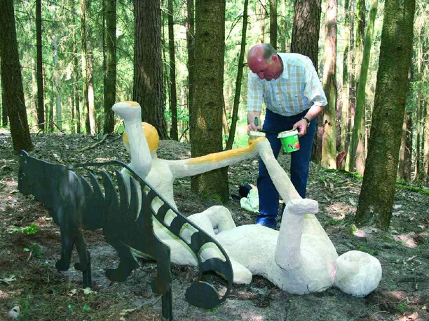 Wilhelm Wortberg Ein Mann bemalt eine liegende Skulptur im Wald, daneben steht eine Metallfigur mit Streifen.