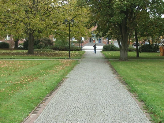 Sedanplatz Ein gepflasterter Weg führt durch einen grünen Park mit Bäumen, im Hintergrund ein Gebäude.