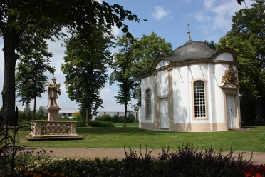 Johanneskapelle Rietberg Kleine weiße Kapelle mit gewölbtem Dach umgeben von gepflegtem Rasen und Bäumen. Sculpture nearby.
