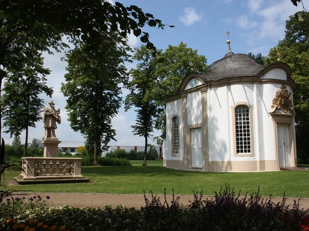 Johanneskapelle Rietberg Kleine weiße Kapelle mit gewölbtem Dach umgeben von gepflegtem Rasen und Bäumen. Sculpture nearby.