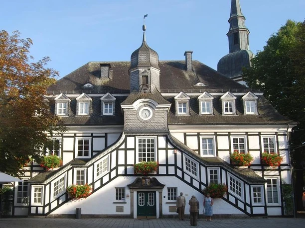 Historisches Fachwerk-Rathaus mit markantem Turm und blühenden Blumenkästen, umgeben von Bäumen.