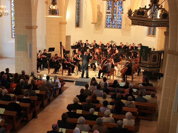 Kirchenkonzert mit Orchester und Publikum, gotische Architektur, Buntglasfenster im Hintergrund.
