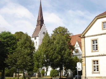 Petri-Kirche Dorfplatz mit Brunnen und Bäumen, im Hintergrund eine weiße Kirche mit spitzem Turm.