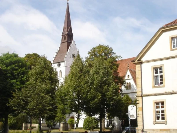 Petri-Kirche Dorfplatz mit Brunnen und Bäumen, im Hintergrund eine weiße Kirche mit spitzem Turm.