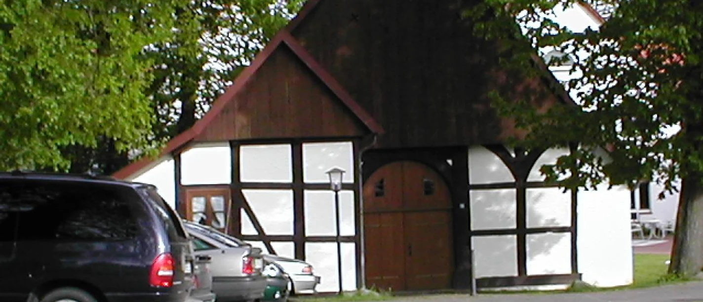 Fachwerkhaus mit weiß verputzten Wänden und dunklem Fachwerk, umgeben von Bäumen und Autos.