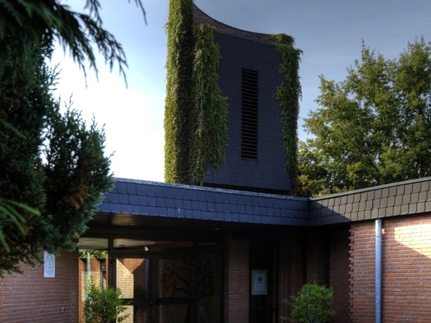 Ev. Versöhnungskirche 3 Moderner Backsteinbau mit begrüntem Turm und Eingang im Schatten hoher Bäume, ruhige Umgebung.