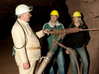 Besucherbergwerk Kleinenbremen Ein Bergwerksführer erklärt zwei Besuchern in Schutzhelmen ein altes, rostiges Bohrgerät unter Tage.