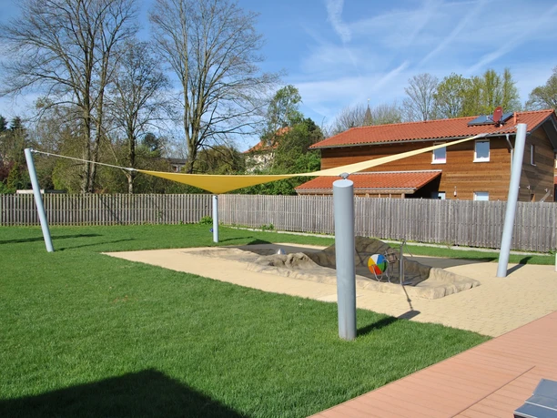 Freibad Spielplatz mit Sandkasten, Sonnensegel und Spielgerät auf grüner Wiese, umgeben von Holzhäusern.
