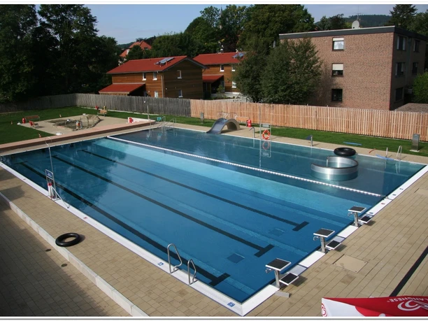 Freibad Ein großes, rechteckiges Freibad mit klar blauem Wasser, umgeben von Bäumen und roten Dächern.