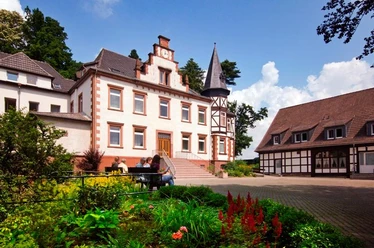 Eine Klosteranlage mit historischem Hauptgebäude und Fachwerkhaus im Grünen, strahlender Himmel.