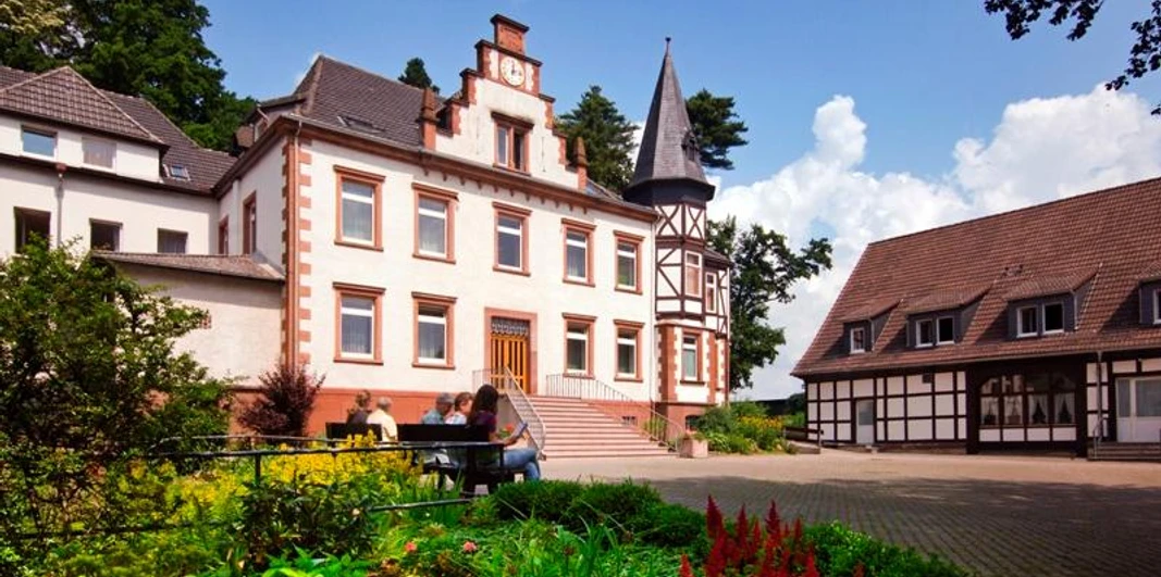 Serviam Schwestern von Warburg-Germete Eine Klosteranlage mit historischem Hauptgebäude und Fachwerkhaus im Grünen, strahlender Himmel.