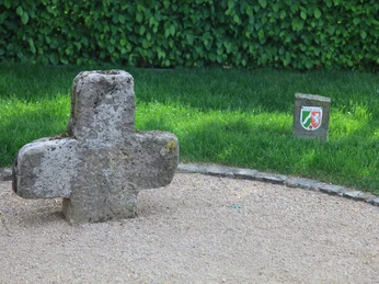 Steinkreuz Versmold Steinernes Sühnekreuz in einer Kiesfläche vor einer grünen Hecke, dahinter ein kleiner Wappenstein.