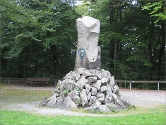 Steinerne Gedenksäule im Wald zu Ehren von Otto von Bismarck, umgeben von Bäumen und Kiesweg.