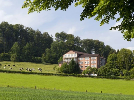 St. Clemens Bad Driburg Das Bild zeigt ein mehrstöckiges Gebäude auf einer grünen Wiese, umgeben von Bäumen und Kühen.