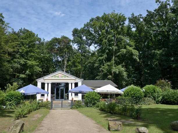 Ein charmantes Café mit klassischem Säuleneingang, umgeben von üppigem Grün und mehreren Sonnenschirmen.