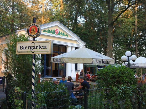 Ein Biergarten im Grünen mit weißen Sonnenschirmen und einem Gebäude im Hintergrund bei Tageslicht.