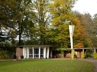 Runder Pavillon mit Glasfront im Grünen, umgeben von hohen Bäumen, mit markantem Fahnenmast davor.