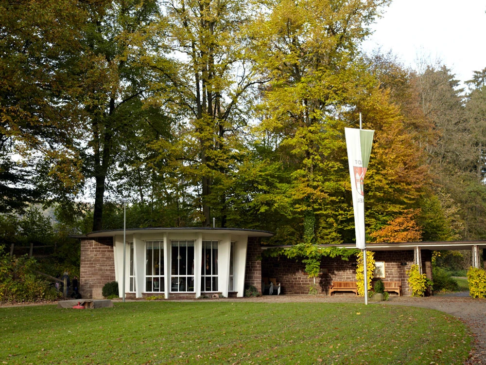Kaiserbrunnen Brakel Runder Pavillon mit Glasfront im Grünen, umgeben von hohen Bäumen, mit markantem Fahnenmast davor.