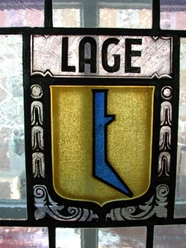 Wappen im Haus Grotebrune Buntes Bleiglasfenster mit einem Wappen, das ein abstraktes, blaues Symbol auf goldenen Hintergrund zeigt.
