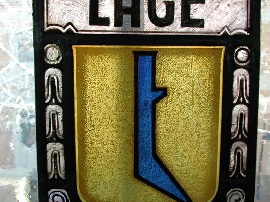 Wappen im Haus Grotebrune Buntes Bleiglasfenster mit einem Wappen, das ein abstraktes, blaues Symbol auf goldenen Hintergrund zeigt.