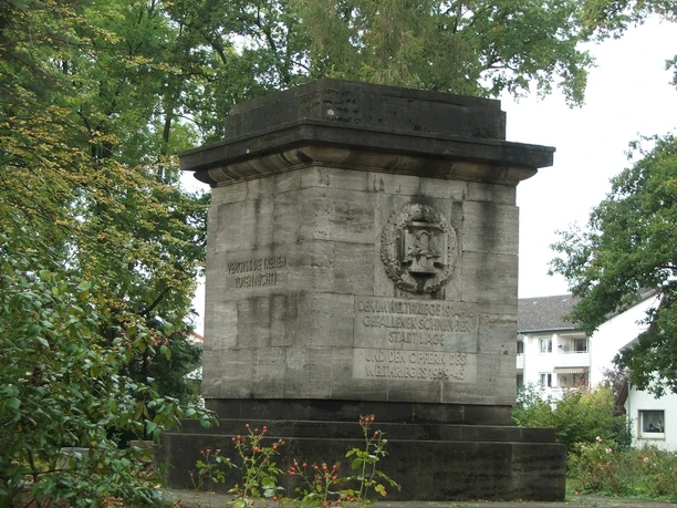 Gedenkstein aus Stein mit Inschrift, umgeben von Bäumen im Friedenspark, erinnert an historische Ereignisse.